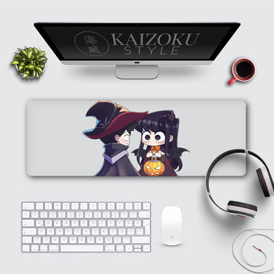 A desk mat featuring chibi-style Komi San from Komi Can't Communicate in a cute Halloween theme. She is dressed as a bat, holding a pumpkin bucket, while another character in a witch hat looks at her. The design is lighthearted and festive, adding charm to the workspace.